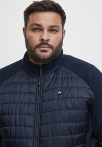 Jūrų jėgos pūkinė striukė su tarde viršutine dalimi, vientisa apatine dalimi ir rankovėmis, stovinčiu apykakle, priekiniais užtrauktukais ir mažuoju logotipu kairėje krūtinės pusėje.