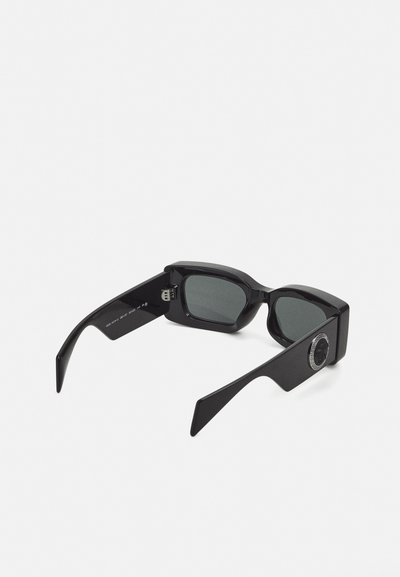 Lunettes de soleil noires avec une monture carrée, verres foncés et un emblème rond texturé sur le côté. Matière légère et finition brillante.