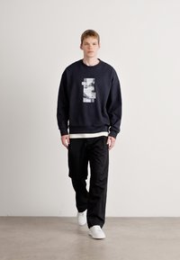 Felpa blu navale con un design grafico frontale, abbinata a pantaloni neri e sneakers bianche, presentata in una posa eretta su uno sfondo neutro.