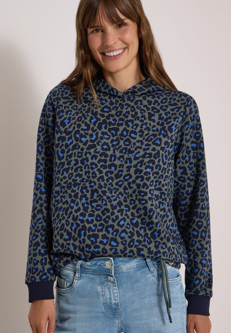 Femme souriante portant un sweat à capuche bleu et noir à imprimé léopard et un jean en denim bleu clair sur un fond clair uni.
