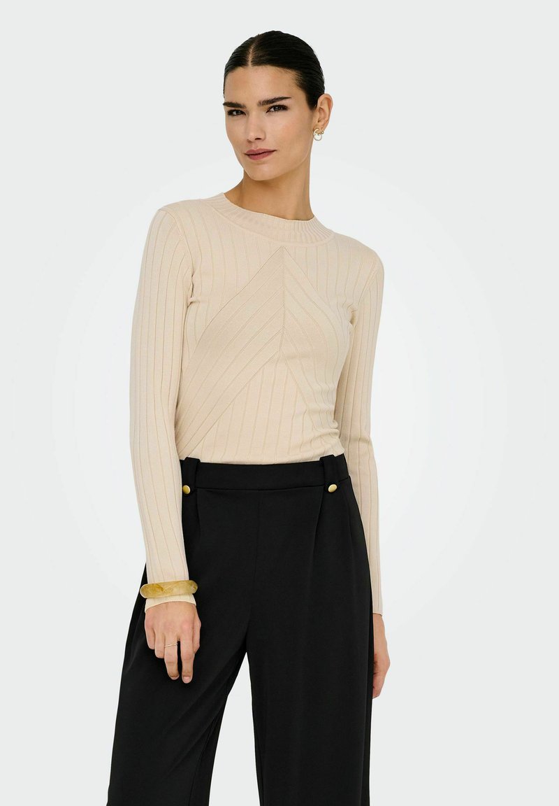 Kvinna med släta mörka hår som bär en beige ribbad långärmad topp och högmidjade svarta byxor med guldknappar, accessoriserad med ett gult armband.
