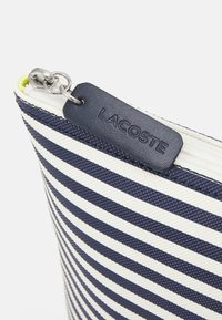 Pochette rayée bleu marine et blanche avec une surface texturée ; dotée d'une fermeture éclair en argent et d'une étiquette logo Lacoste en faux cuir bleu marine.