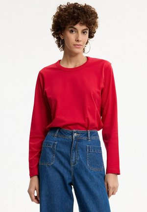 Mujer con cabello rizado que lleva pendientes grandes de aro, una camiseta roja de manga larga y vaqueros azules de talle alto sobre un fondo liso.
