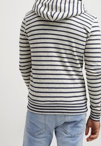 Felpa a righe in tessuto crema e blu navy, con cappuccio e maniche lunghe. La texture sembra morbida; il motivo è costituito da linee orizzontali distanziate uniformemente.
