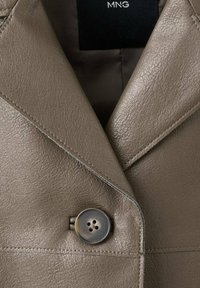 Kožená bunda v tóne taupe s golierom s rozdeleným klopou a jedným tmavým zapínaním na gombík, zdobená viditeľnými švami.