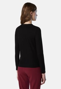 Suéter negro acanalado con cuello redondo y mangas largas, combinado con pantalones de color rojo oscuro. La tela texturizada crea una silueta ajustada.