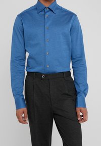 Camicia blu a maniche lunghe in tessuto morbido con collo classico e bottoni in legno, abbinata a pantaloni grigio scuro con piega piatta.