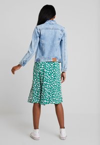 Lichtblauwe spijkerjack met een aansluitend ontwerp, heeft lange mouwen en een label op de rug. Gepaard met een groen rok met bloemenpatroon.