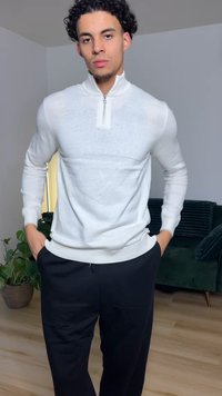 White zip-up sweater with a high collar, ribbed cuffs, and hem, paired with black pants. Background features a green couch and plant.