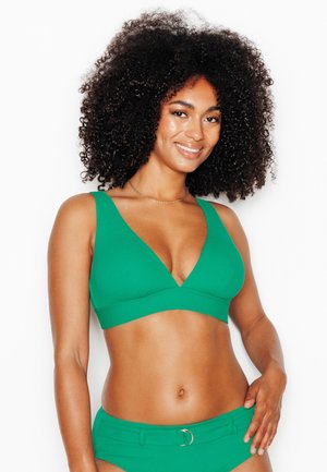 Femme aux cheveux bouclés portant un maillot de bain deux pièces vert, souriante, debout sur un fond blanc, main posée sur la hanche.
