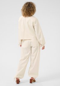 Lichtbeige katoenen jas en broekset, met een cropped ontwerp, elastische manchetten en trekkoorden aan de enkels. Vierkante blokhakken maken de look compleet.