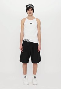 Hvit singlet, svarte shorts med elastisk midje, og en svart lue med hvit tekst. Skoene er hvite med merke detaljer.