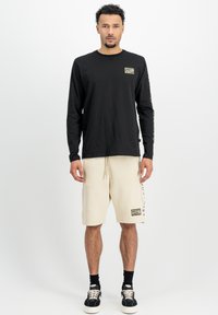 Camiseta negra de manga larga con un pequeño parche, combinada con pantalones cortos crema que cuentan con texto lateral y un parche, llevada con zapatillas negras y calcetines.