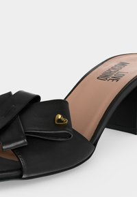 Μαύρα ψηλοτάκουνα mules με ανοιχτή μύτη, ευρύ διπλωμένο λουράκι από δέρμα με μικρή χρυσή καρδιά και μπρονζέ εσωτερική σόλα με το λογότυπο «Love Moschino».