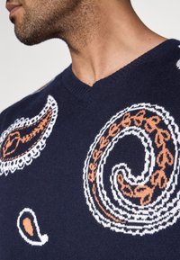 V formos iškirpte mezgimo tamsiai mėlynas megztinis su paisley raštais. Savybės balti ir oranžiniai detalūs akcentai. Minkšta tekstūra su ribotu kaklo apvadu.