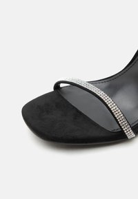 Μαύρο σανδάλι ανοιχτού δαχτύλου με λεπτό λουράκι διακοσμημένο με μικρές λαμπερές πέτρες ριντόν στο μπροστινό μέρος.