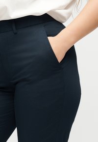 Χέρι στην μπροστινή τσέπη σκουρόχρωμων κολλητών παντελονιών που φοριούνται με λευκό τοπ σε απλό φόντο.