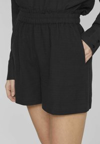 Juodi šortai, pagaminti iš tekstūruotos medžiagos, turi elastingą juosmenį ir šonines kišenes. Dizainas yra laisvas, su apdirbtais kraštais.