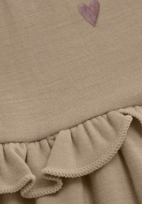 Beige stickad plagg med en horisontell volang i botten och en liten lila hjärta brodering. Mjuk textur, enkel design.