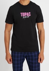 Černé bavlněné tričko s růžovým grafickým textem "TUPAC", krátké rukávy a kulatý výstřih. V kombinaci s modrými kostkovanými kalhotami.