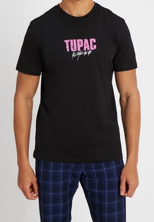 Camiseta de algodón negra con gráfico de texto rosa "TUPAC", mangas cortas y cuello redondo. Combinada con pantalones de cuadros azules.