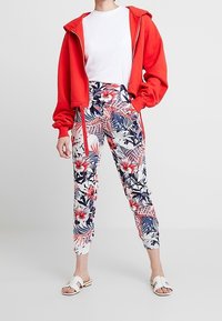 Haut zippé rouge, t-shirt blanc et jogging à imprimé floral avec des fleurs rouges et bleues sur fond blanc, associé à des sandales blanches.