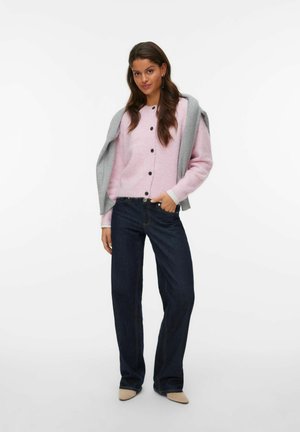 Cardigan bouton rose clair superposé sur un pull gris, associé à un jean à larges jambes bleu foncé. Détails notables : boutons noirs et texture légère en tricot.