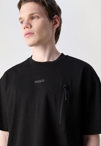 Černé tričko s kulatým výstřihem, s kapsou na zip na levé straně a reliéfním logem "HUGO" nahoře. Hladká bavlněná tkanina.