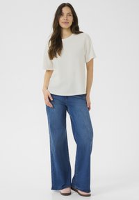 Vit kortärmad topp i kombination med högmidjade vida blå jeans och ljusa skor, på en enfärgad bakgrund.