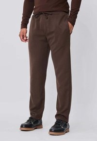 Homme portant un pantalon décontracté marron avec taille à cordon et des chaussures noires à lacets avec semelles épaisses marron, debout avec une main dans la poche.