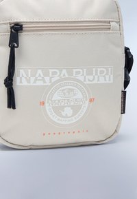 Beige fabric shoulder bag featuring a front zip pocket, printed logo, and a textured surface with black accents and a pull cord.