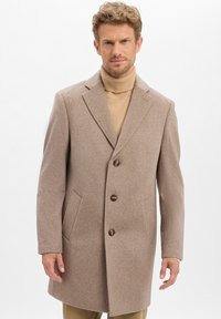 Mann, der einen beigen Wollmantel über einem hellbraunen Rollkragenpullover trägt, vor einem schlichten weißen Hintergrund steht.