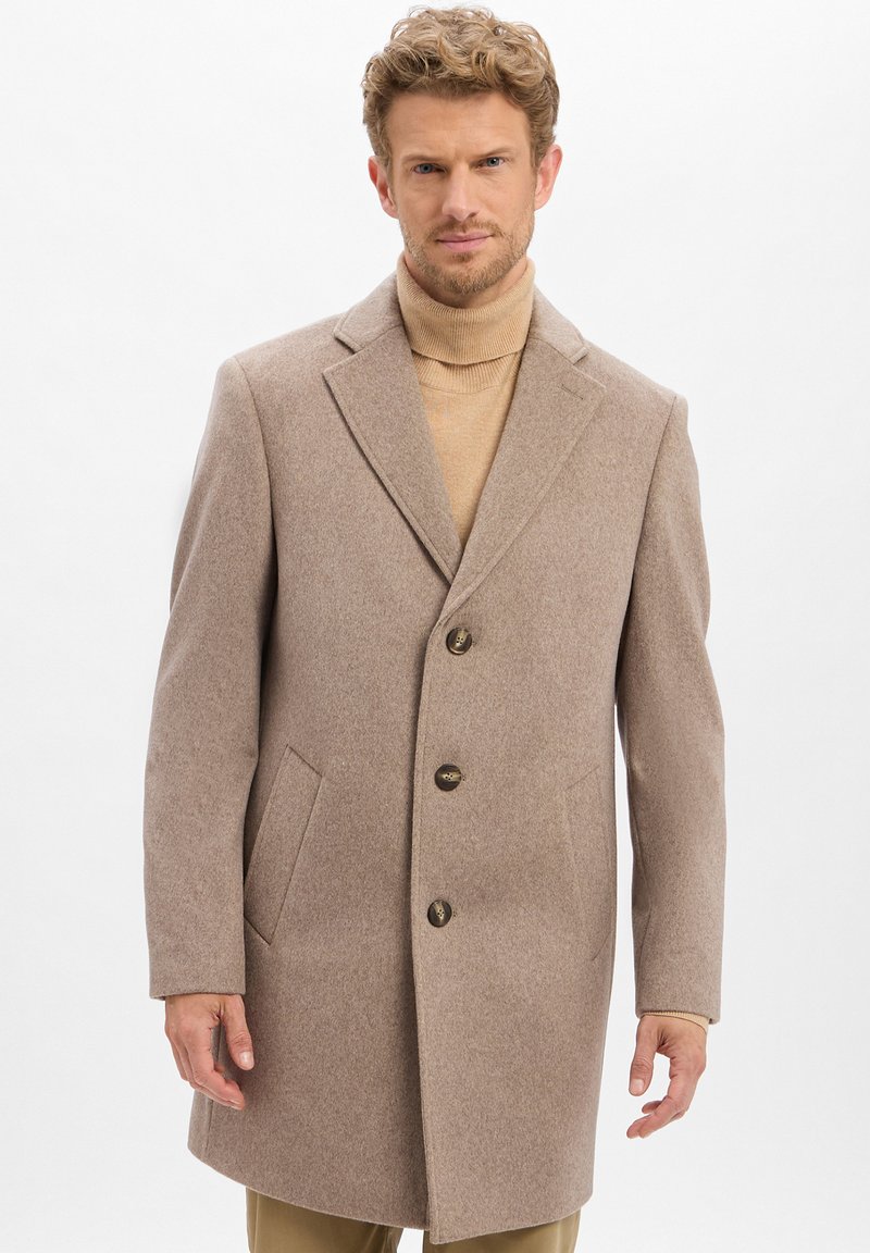Mann, der einen beigen Wollmantel über einem hellbraunen Rollkragenpullover trägt, vor einem schlichten weißen Hintergrund steht.