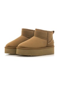 Botas de ante marrón hasta el tobillo con un forro suave y peludo, con punta redonda y una suela plana y texturizada. Costura decorativa en el lateral.