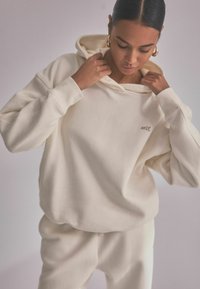 Cream-colored oversized hoodie made of soft fabric, featuring a front pocket and the word "self." embroidered on the chest in small letters.