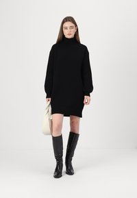 Juoda raukšlėta megzta turtlenecko suknelė ilgomis rankovėmis, derinama su juodais iki kelių batais ir kreminės spalvos rankine. Paprasta, kūną apglėbianti siluetas.