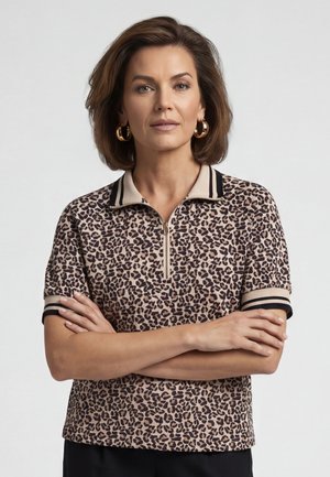 Vrouw met kort bruin haar en gouden oorringen in ringvorm, draagt een beige en zwart short-sleeve shirt met luipaardprint en een ritskraag, armen gekruist.