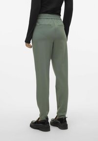 Groene elastische broek met een gladde textuur, slim fit, achterzakken, gecombineerd met zwarte instap schoenen op een witte achtergrond.