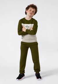 Chlapec s kučeravými vlasmi stojí so zloženými rukami, oblečený v zeleno-beige mikine Champion, zodpovedajúcich nohaviciach a čiernych teniskách na bielom pozadí.