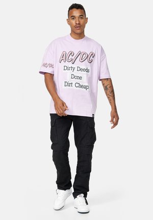 Camiseta gráfica de AC/DC en rosa claro con texto negro, combinada con pantalones cargo negros y zapatillas blancas. La camiseta tiene un corte oversize y una impresión en negrita.