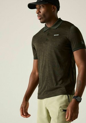 Hombre con gorra negra, polo de manga corta verde oscuro con logo, pantalones beige y reloj de pulsera negro, de pie frente a un fondo liso.