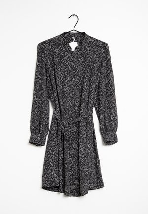 Robe noire à manches longues avec des pois blancs, ceinture à la taille cintrée et poignets boutonnés, suspendue sur un cintre noir sur fond blanc.