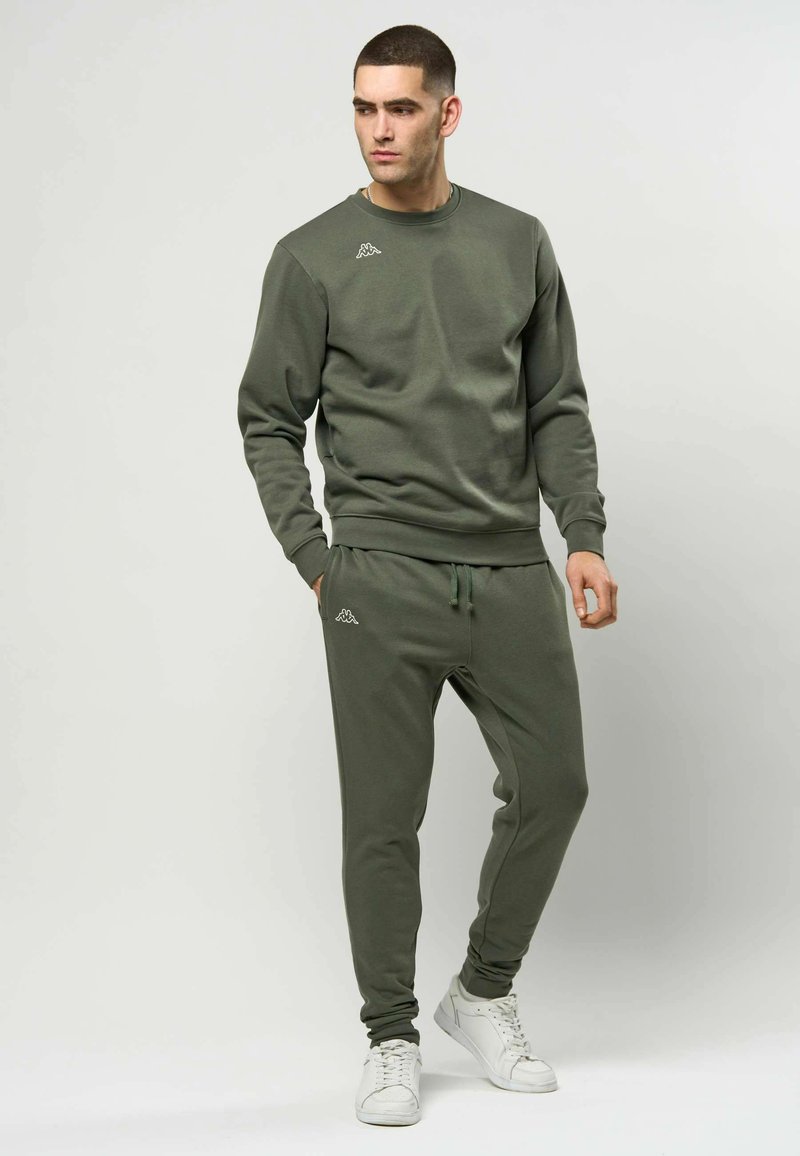 Olivengrønt sett med genser og joggebukser laget av mykt stoff. Genseren har rund hals og subtil logo. Joggebuksene har elastisk midjebånd.