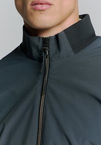 Donkergrijze ritsjack met een hoge kraag, gladde stof en een metalen rits met een gestructureerd accent. Close-up van de halslijn.