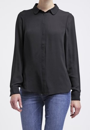 Frau trägt eine schwarze langärmelige Bluse mit Kragen und blaue Jeans, steht vor einem schlichten hellen Hintergrund.