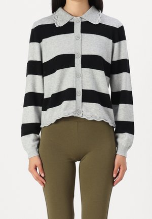 Kvinde iført en grå og sort bredstribet cardigan med knapper, bølget krave og kant, kombineret med olivengrønne tætsiddende bukser.