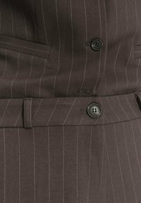 Traje marrón con rayas, que presenta un diseño de un solo botón, botones negros y bolsillos laterales. La tela tiene una textura suave con rayas blancas contrastantes.