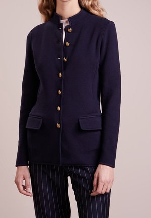 Mujer con chaqueta azul marino ajustada con botones dorados y bolsillos con solapa, combinada con pantalones azul marino de rayas finas sobre un fondo neutro.