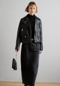 Veste de motard en cuir noir avec des zips et des poches argentées, assortie à un col roulé fitted noir et une longue jupe en denim. Sac à main noir avec chaîne.