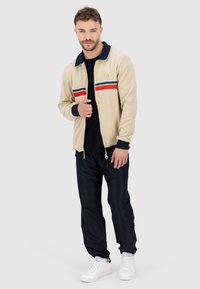 Beige Reißverschlussjacke mit marinefarbenem Kragen und Streifen in Rot, Grün und Weiß, kombiniert mit schwarzen Hosen und weißen Sneakers. Weicher Stoff, legere Passform.